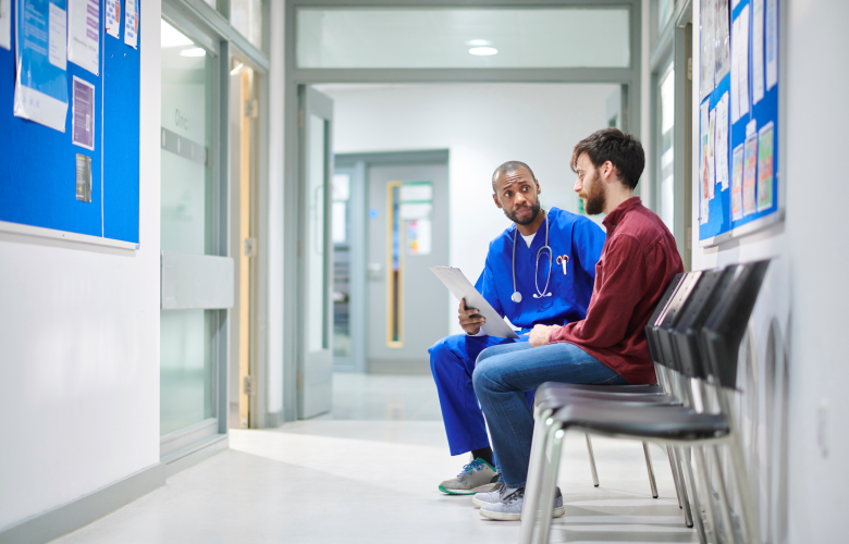 A male doctor sat in a hospital corridor talking with a male patient in his twenties.