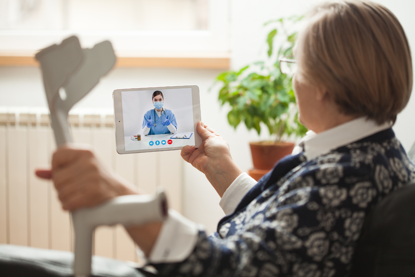An elderly lady, sat down, is holding a walking crutch in one hand and holding a computer tablet in the other hand, having an online appointment with her GP.