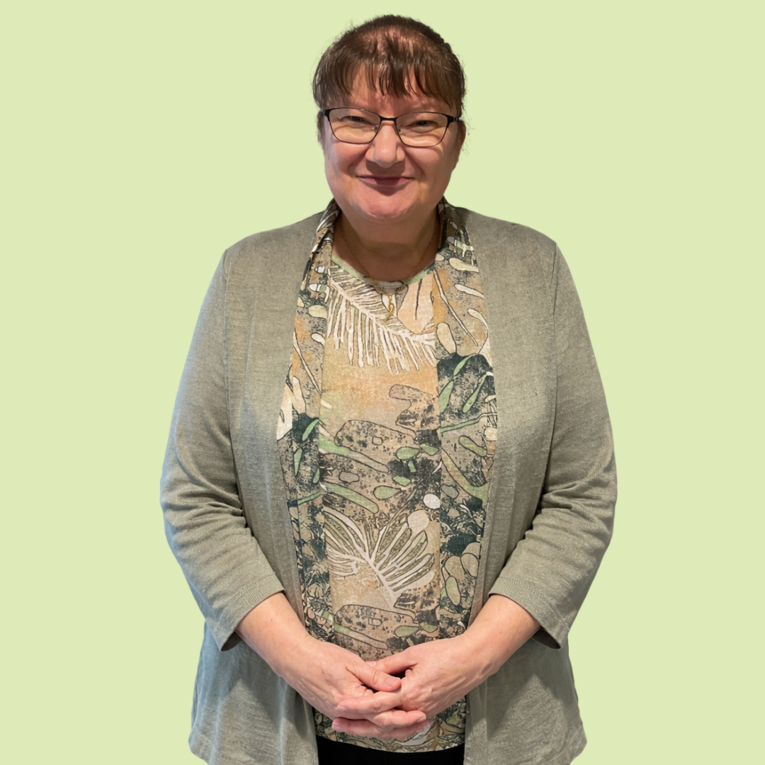 A woman with short brown hair and glasses stands against a plain pale green background, smiling gently at the camera. She is wearing a light sage green cardigan over a patterned blouse in beige and green tones, with black trousers. Her hands are loosely clasped in front of her.