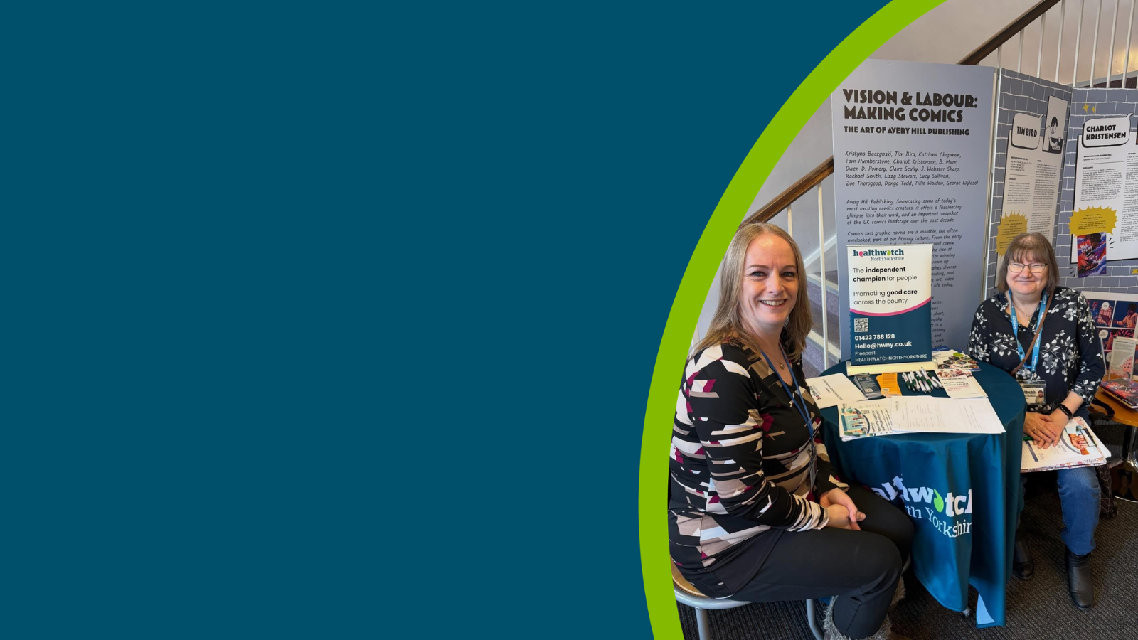 Two women sit smiling at a round table with a teal Healthwatch North Yorkshire tablecloth. The background shows a staircase with white railings and a wooden handrail.