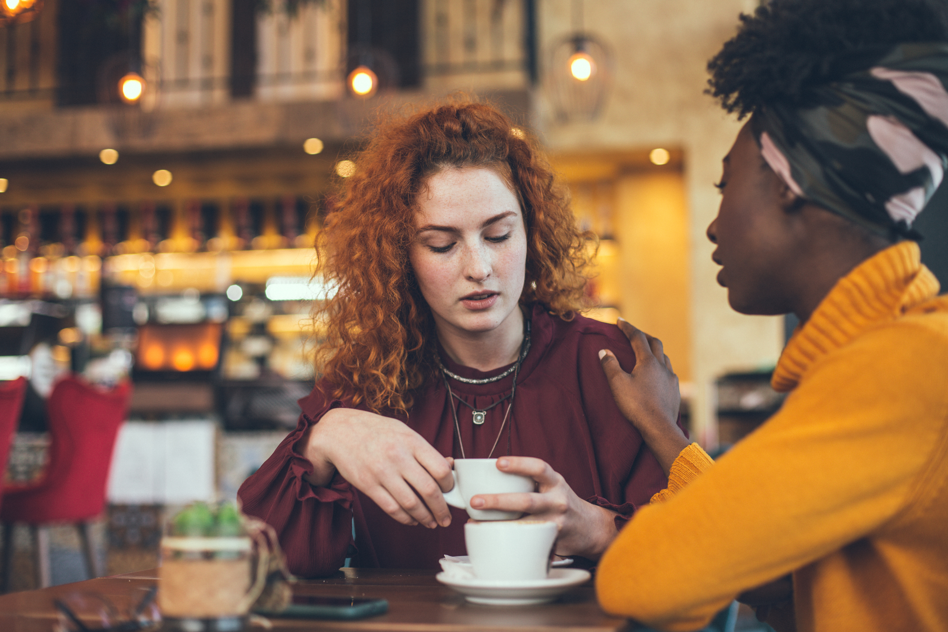 Two female friends in a coffee shop, one consoling the other as they talk