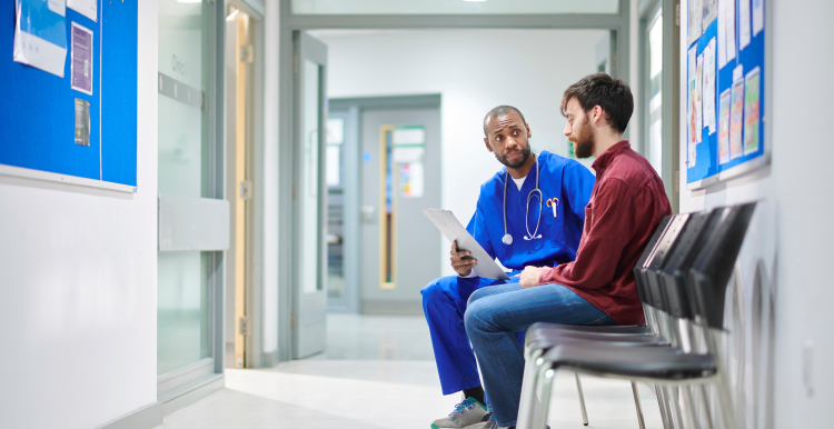 A male doctor sat in a hospital corridor talking with a male patient in his twenties.