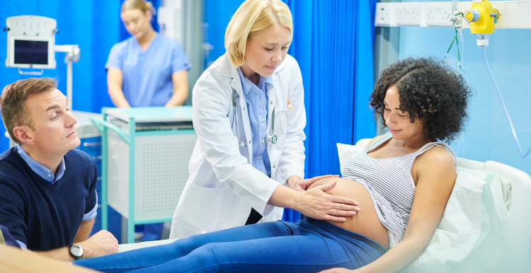 A young pregnant woman laid on a hospital bed, with a female doctor examining her stomach with the woman's husband sat at the bedside.