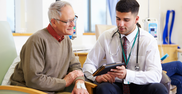 A young male doctor sat in a consultation with an older patient, looking at a mobile tablet