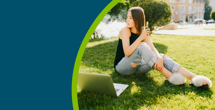 A young person sat on some grass, next to her laptop, using a disposable vape.