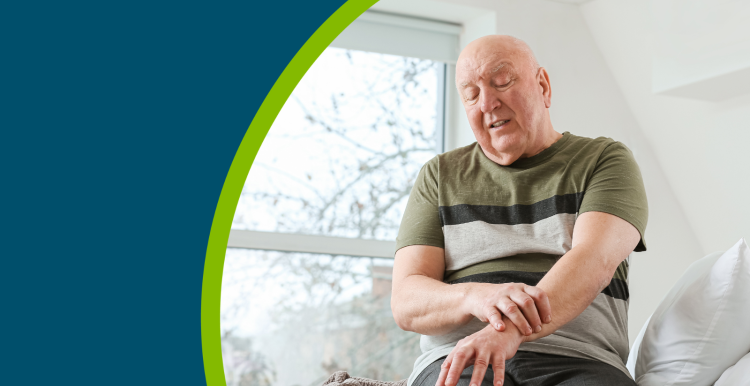 An older man with Parkinson's sat on his bed and holding his wrist.