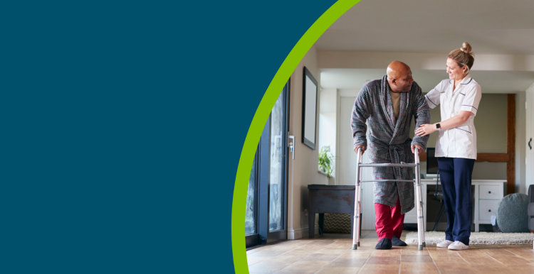 Older man in dressing gown using walking frame being helped by female care worker.