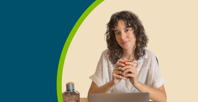 A young female holding a cup of tea and sat at a desk