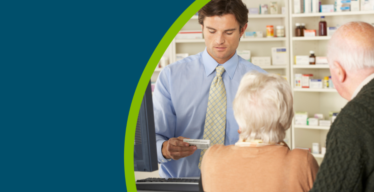 Male pharmacist in pharmacy holding a box of tablets whilst serving an older couple at the counter.and looking at his computer.