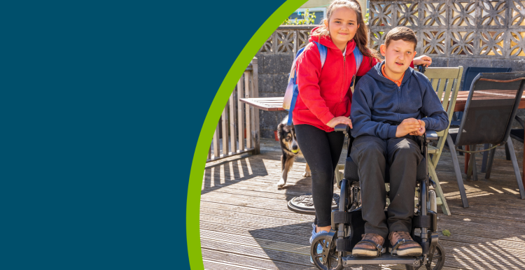 A young female carer with a young person in a wheel chair. Both are smiling to the camera outside on some decking on a sunny day.
