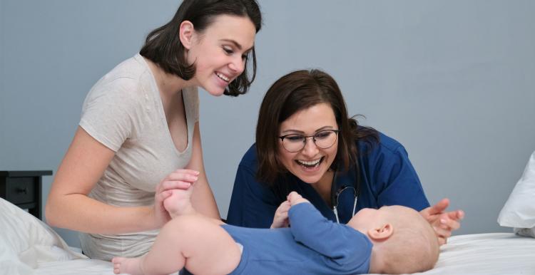 Pediatrician woman doctor in blue uniform with stethoscope talking to young mother of little boy, examining 7 month old baby at home