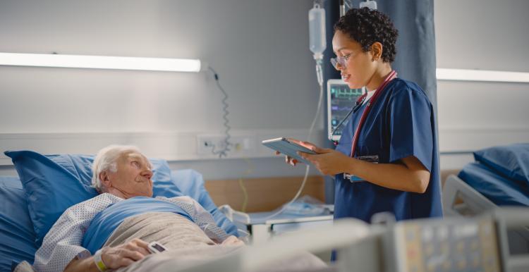 Friendly Female Head Nurse Making Rounds does Checkup on Elderly Patient Resting in Bed.  She Checks Computer for Vitals while Old Man Fully Recovering after Successful Surgery.