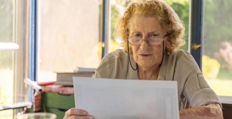 Older woman wearing eyeglasses, reading documents while sitting at a table at home.