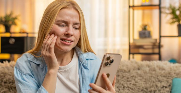 Young woman feeling tooth pain at home, worried face, searching phone for dentist