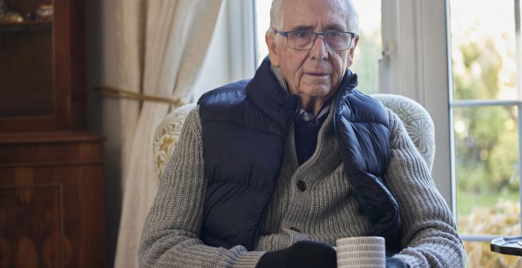 Older man wearing extra clothes with hot drink trying to keep warm at home in an energy crisis