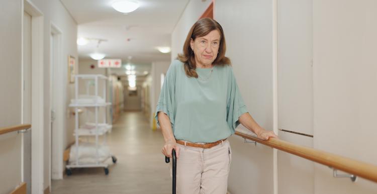 Older woman with a disability holding a railing on a hospital corridor whilst waiting for care.