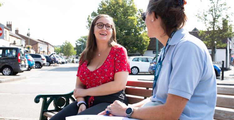 Two women sit on a bench in a town centre on a sunny day, talking and smiling. One woman in a red patterned top faces the other, who is wearing a light blue polo shirt and lanyard and holding a clipboard with papers. Parked cars, buildings and trees are visible in the background.