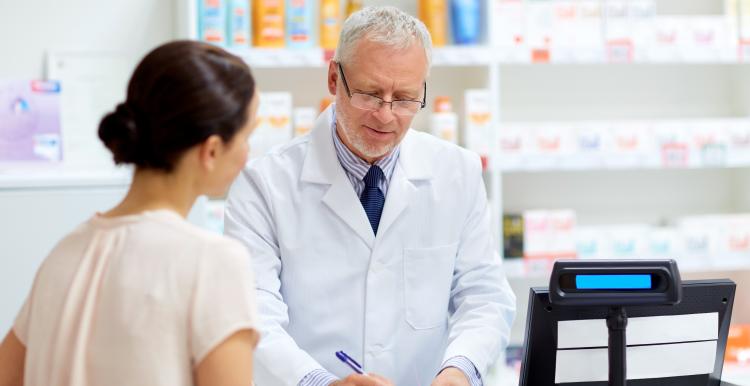 A pharmacist in a white coat stands behind a pharmacy counter, writing on a prescription form while speaking with a woman on the other side. Shelves of medicines and health products are visible in the background.