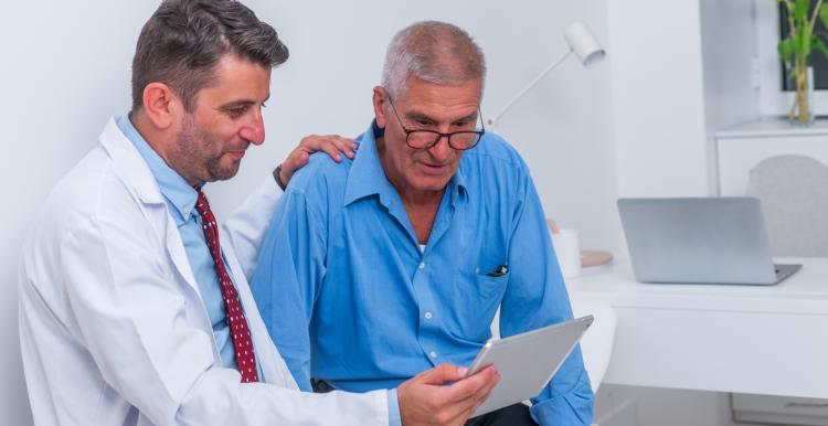 Doctor in a white coat sits beside an older man in a blue shirt, resting a reassuring hand on his shoulder while showing him information on a tablet. They are in a bright clinical room with a desk, laptop, and sunflowers in the background.