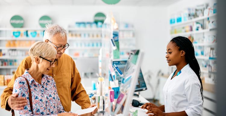 An older couple stand at the counter of a bright community pharmacy, speaking with a pharmacist in a white uniform. The woman holds a prescription slip while the man stands beside her with his arm around her shoulder. Shelves of medicines and health products are visible in the background.