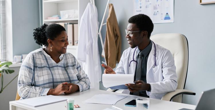 Doctor sitting at a desk in a GP surgery smiles and speaks with a woman during a consultation, holding paperwork. The room contains medical posters, shelves, a stethoscope around the doctor’s neck, and a laptop on the desk.