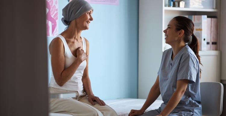 Middle aged woman with headscarf sitting on examination table, smiling and talking with young adult female nurse, in medical clinic with breast cancer awareness posters visible