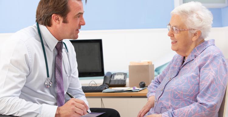 Doctor talking to an older female patient in doctor's surgery. He is writing a prescription and she is smiling whilst looking at him.