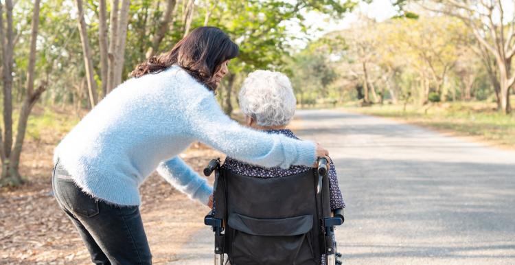 An unpaid carer pushing an older person in a wheelchair along a peaceful tree-lined path.