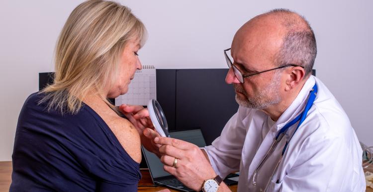 A GP examines a patient's shoulder with a handheld magnifying device during an appointment in a GP consultation room.