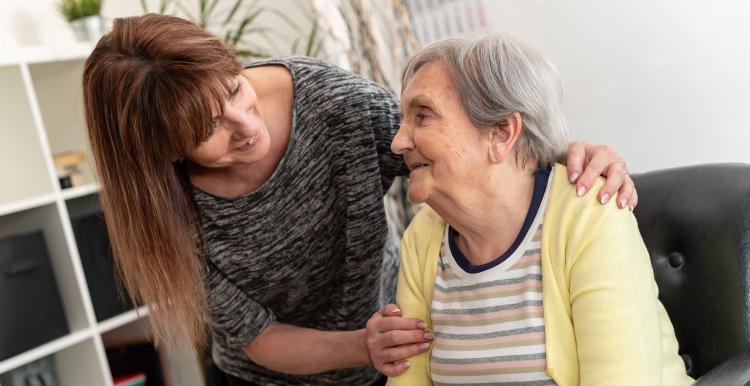 A woman places a reassuring hand on the shoulder of an older woman as they smile and make eye contact while talking in a comfortable home setting. The image represents care, support and companionship for older people.