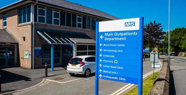 Exterior of an NHS hospital outpatient department on a bright sunny day. A large blue and white NHS direction sign stands in the foreground, pointing towards the Main Outpatients Department, Blood Donor Centre, Dermatology Unit, Endoscopy, Pharmacy, Ashton Road Clinic, Medical Unit 1 and Orthopaedic Clinic. The hospital building and entrance are visible behind the sign, with a car parked outside and a road running alongside the site.