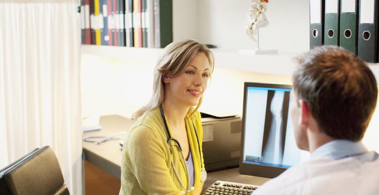 Female healthcare professional wearing a stethoscope talking with a male patient in a GP consultation room.