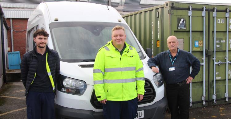 Three members of the hospital transport team stand beside a white minibus outside a hospital service building. The person in the centre is wearing a bright yellow high-visibility jacket, while the two others wear dark work clothing. Large green storage containers and hospital buildings can be seen behind them.
