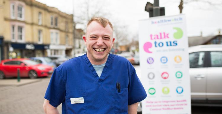 NHS worker standing infront of a Healthwatch sign that says 'talk to us'