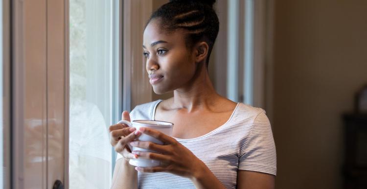 A woman looking sad, looks out of the window holding a cup of tea