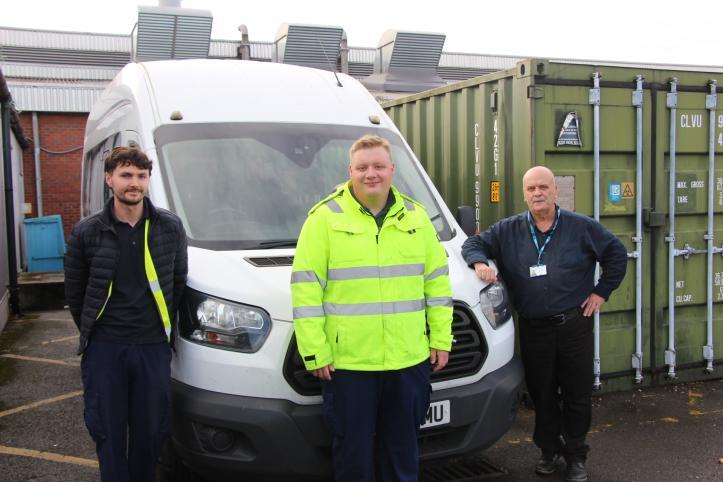 Three members of the hospital transport team stand beside a white minibus outside a hospital service building. The person in the centre is wearing a bright yellow high-visibility jacket, while the two others wear dark work clothing. Large green storage containers and hospital buildings can be seen behind them.