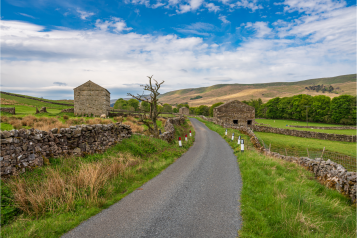 A rural road in Hawes, North Yorkshire - with fields at either side and two stone outbuildings.