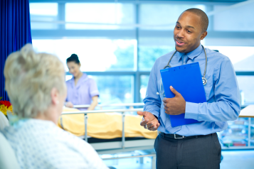 A doctor at the bedside of an elderly female patient holding a clipboard and explaining her treatment to her