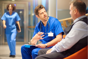 Male doctor talking with an older male patient in a hospital corridor.