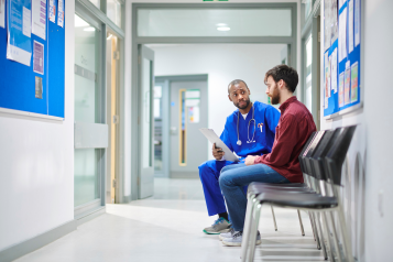 A male doctor sat in a hospital corridor talking with a male patient in his twenties.