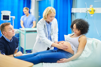 A young pregnant woman laid on a hospital bed, with a female doctor examining her stomach with the woman's husband sat at the bedside.