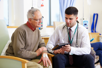 A young male doctor sat in a consultation with an older patient, looking at a mobile tablet