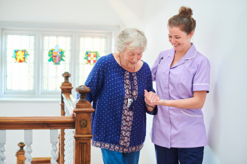 An elderly female resident being helped with her balance at the top of the stairs by a member of care staff