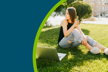 A young person sat on some grass, next to her laptop, using a disposable vape.