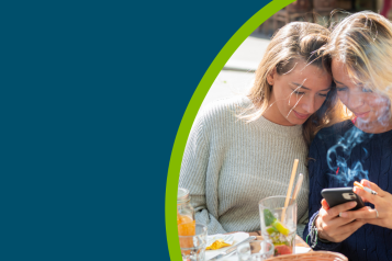 Two young females smoking whilst sat outside a cafe  and looking at one of their smartphones.