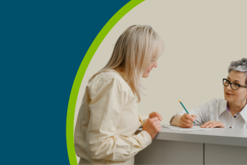 A receptionist at a GP practice' reception desk talking with a patient.