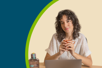 A young female holding a cup of tea and sat at a desk