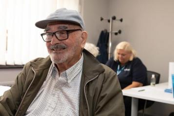 An older man is smiling whilst sat waiting for a healthcare appointment.