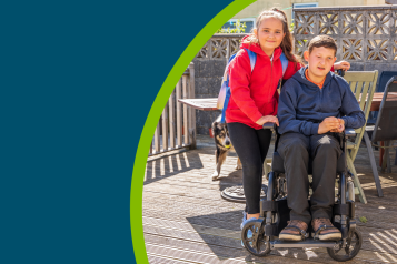 A young female carer with a young person in a wheel chair. Both are smiling to the camera outside on some decking on a sunny day.