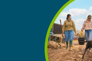 Two Female Farm Workers Walking Across Yard With Dog Past Cattle Barn At Feeding Time